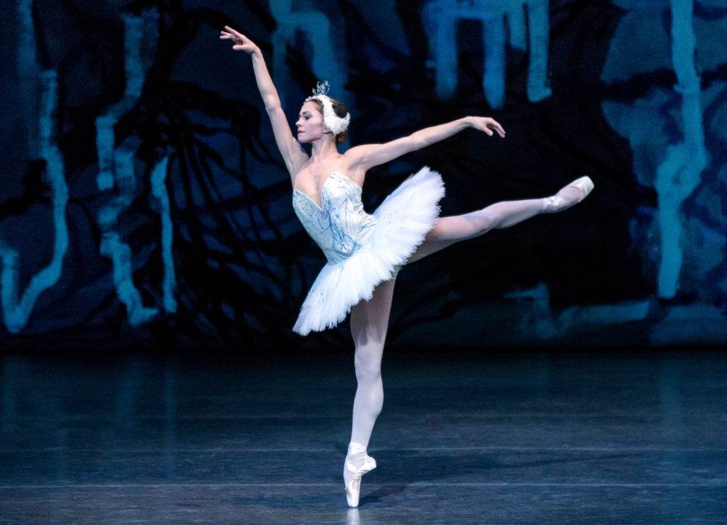 New York City Ballet principal dancer Megan Fairchild holds an arabesque as Odette in Swan Lake. She wears a white tutu and poses against a blue stage backdrop