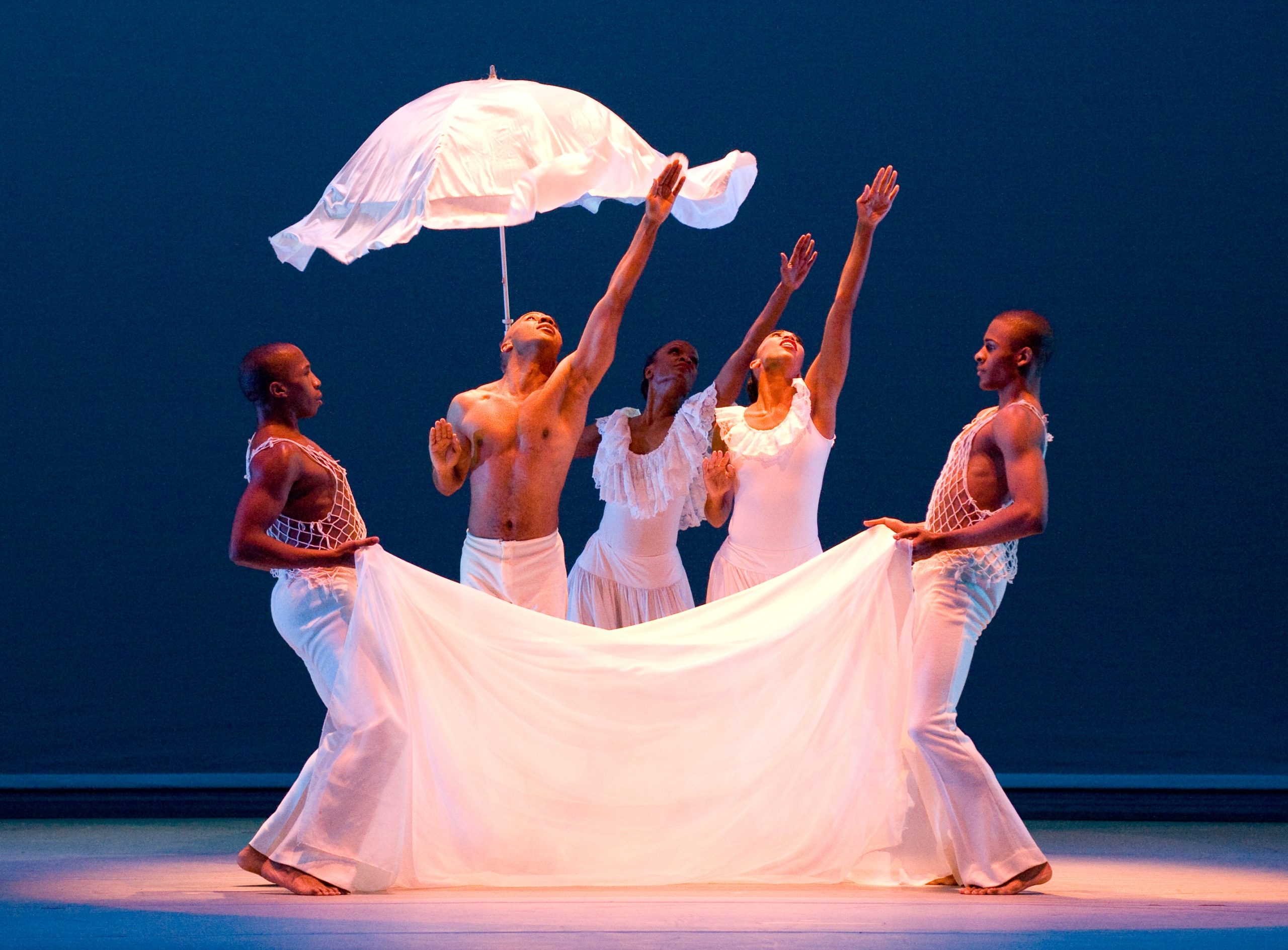 Five dancers stand in a shallow semi circle around a white cloth that they hold up to their legs. One also holds a white cloth umbrella above them. They all look up into the air, and the three middle dancers reach their arms upward with palms facing forward.
