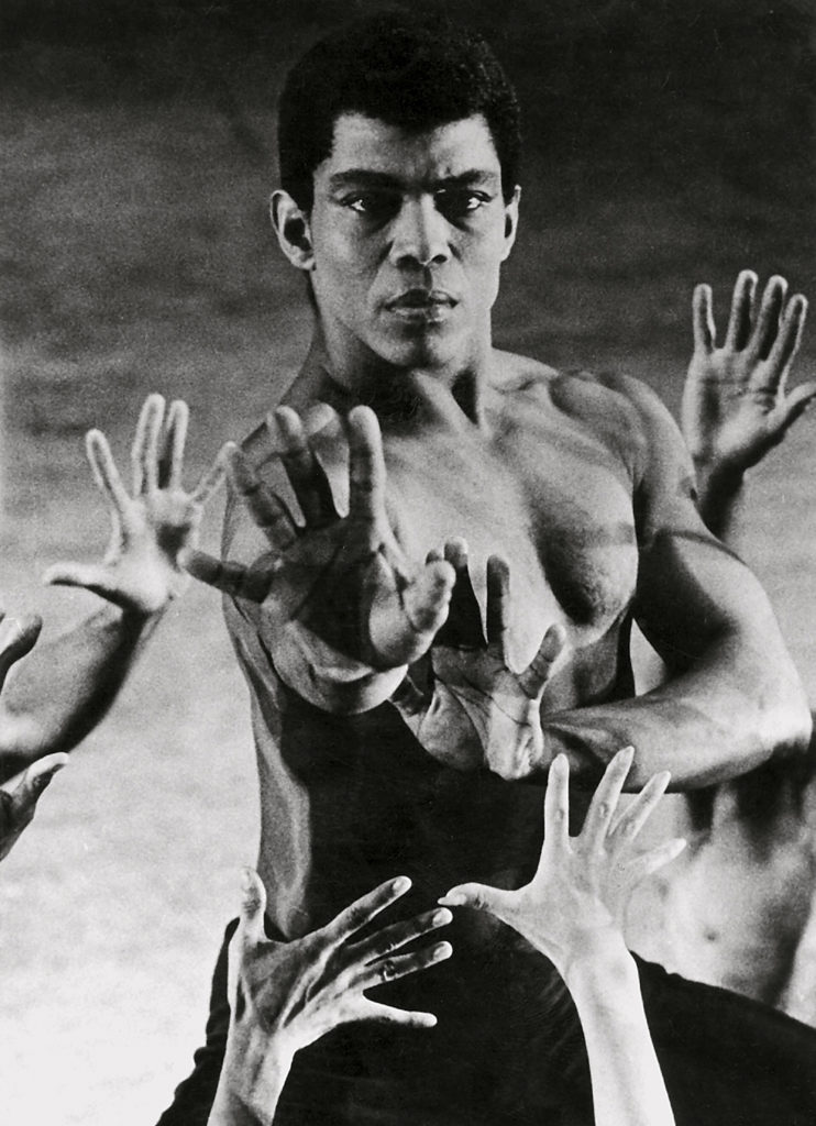 Black and white photo of Alvin Ailey from the torso up, reaching his arms toward the camera with a stern expression. Around him, other hands reach toward him. He wears no shirt and his hair is short around his head.