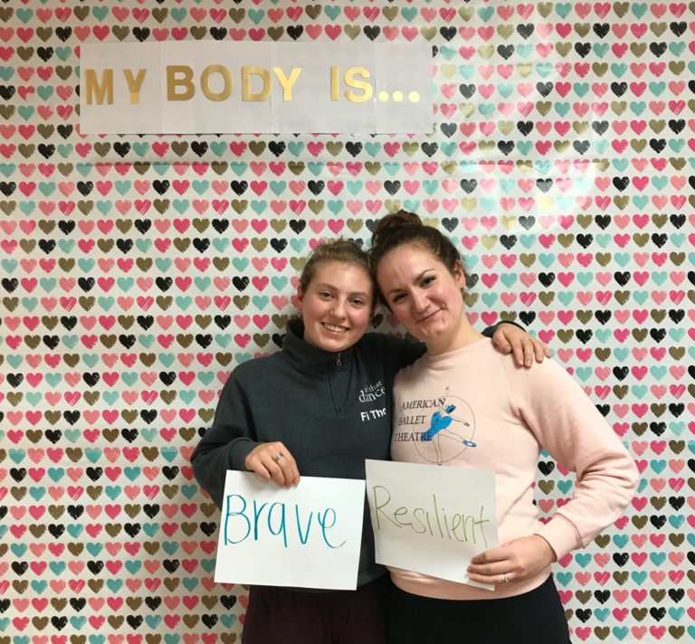 Two dance students standing and holding signs that read 'Brave' and 'Resilient' against a wallpaper filled with heads and a sign that reads 'My Body Is...'