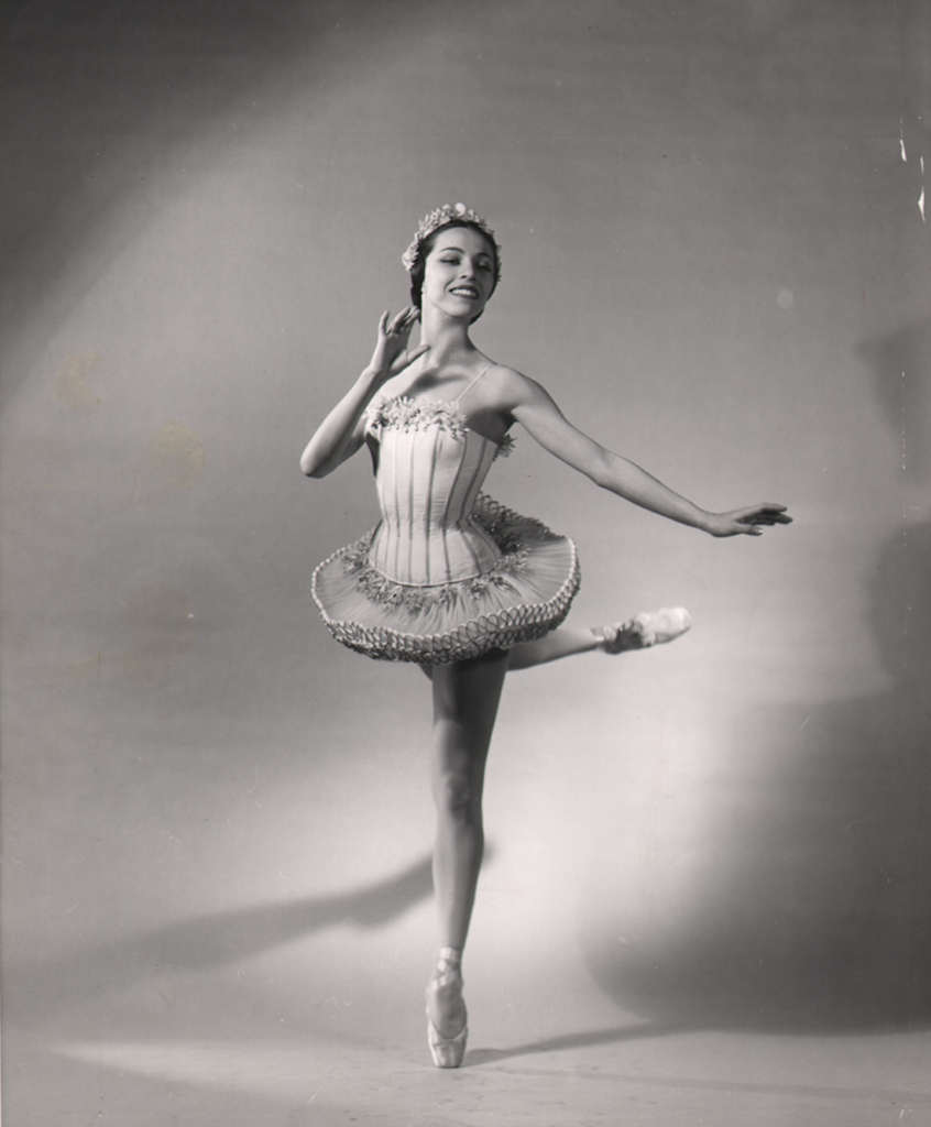 Maria Tallchief poses in attitude derrier as the Sugar Plum Fairy. She wears a short tutu and crown, with one hand held to her face and the other in demi seconde.