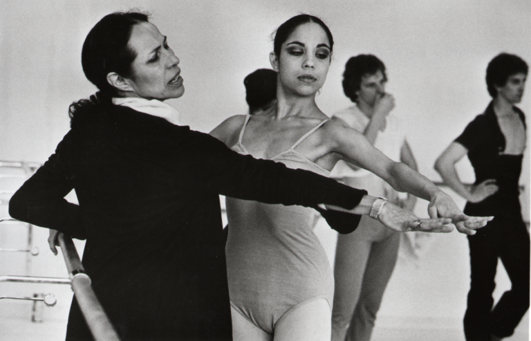 Maria Tallchief teaches class. She stands in front of a female student, mirroring her position with an outstretched front arm. She wears a dark sweater and her hair is pulled back.