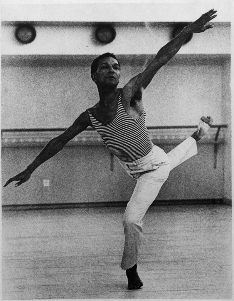 Black and White photo of Talley Beatty in a dance studio. His arms are outstretched on a diagonal with his back leg lifted in attitude behind him. He holds a deep plié on his supporting leg.