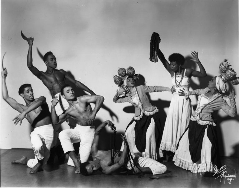 Black and white photo of Tally Beatty and other dancers in a standing tableau pose centering around Beatty who lays on the floor. Men wear light colored pants and no shirt. Women wear long dresses with lace-lined jackets and large headpieces.