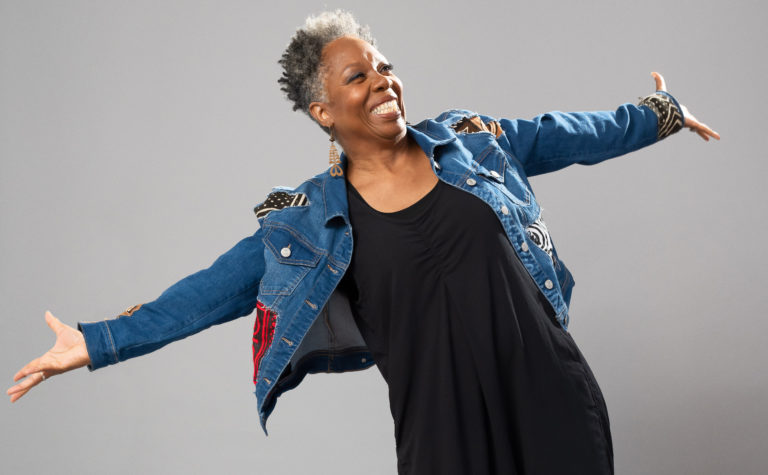 Image of dance artist and educator Jawole Willa Jo Zallar smiling with arms extended. She wears a black dress and denim jacket.