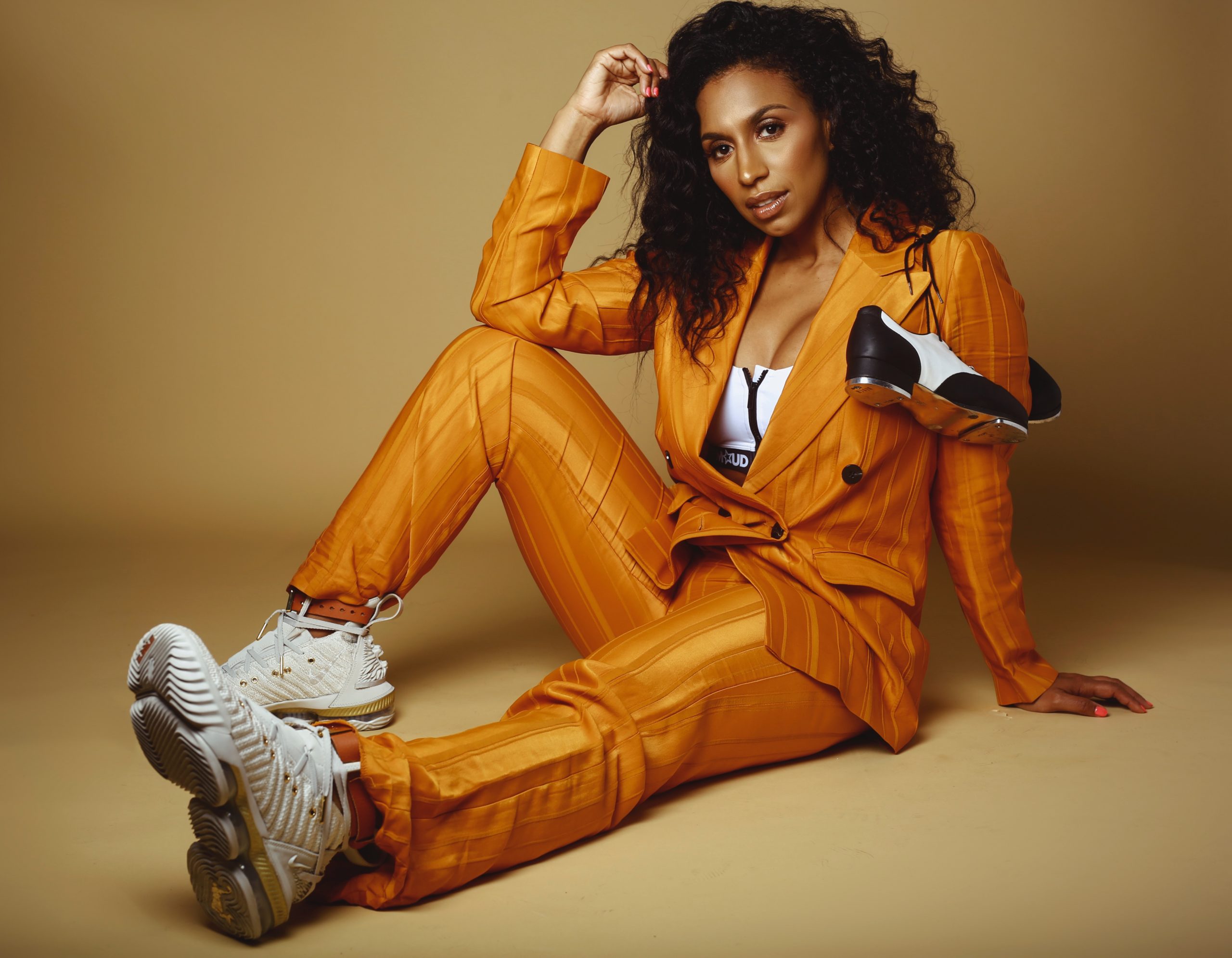 Tap dancer Chloe Arnold in an orange suit sits on the floor with tap shoes across her shoulder