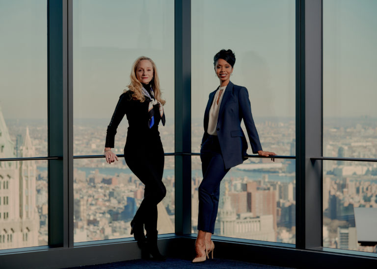 Darla Hoover and Aesha Ash lean against ballet bars set before floor to ceiling windows showing New York City. Darla's blonde hair curls to her shoulders; she wears all black with a colorful scarf. Aesha smiles warmly at the camera, dressed in a blue suit and nude heels.