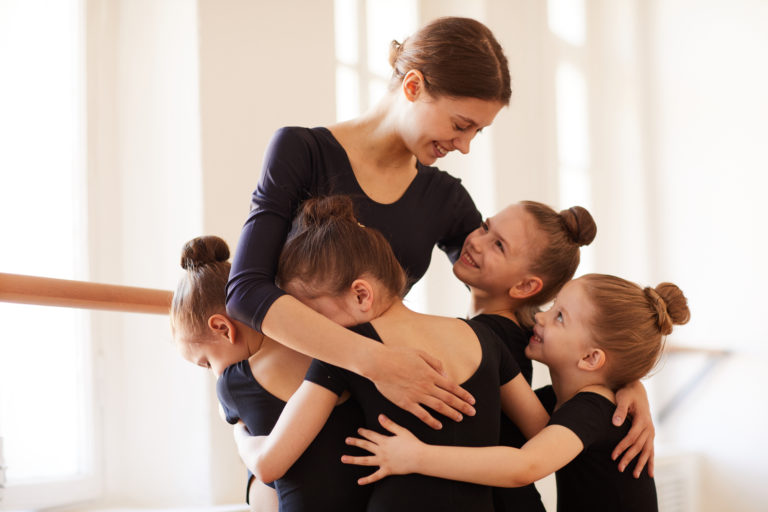 Group of little girls hugging ballet teacher in studio lit by warm sunlight, copy space