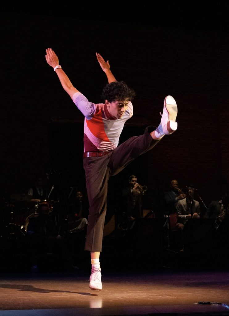 Image of Caleb Teicher on stage with his arms held back and kicking one leg off the ground