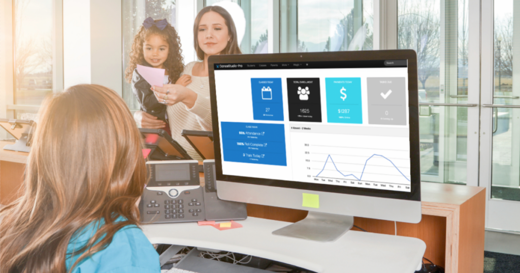 A woman checks her young daughter in for dance class at the reception desk. DanceStudio-Pro's software is open on the computer monitor.