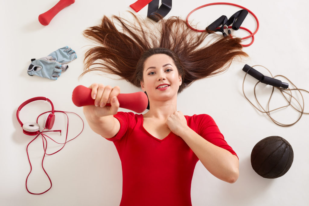 Relaxed sportswoman surrounded with headphones, rolling skip, fitness gloves, elastic bands, expander, medball, lady wearing red outfit laying on white surface, holding red dumbbell in hand.