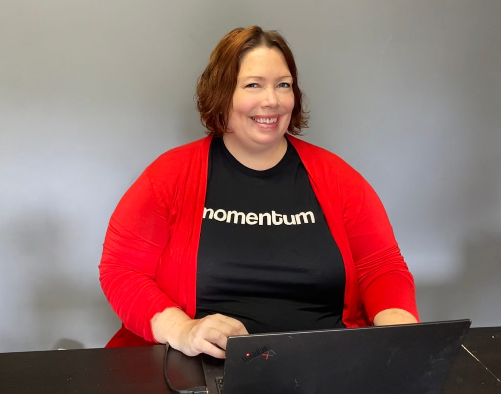 Maggie Walls, in a red sweater and black shirt with her studio's name on it, smiles while standing in front of her laptop.