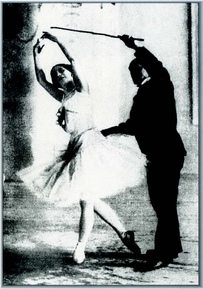A male dance teacher helps a ballerina with her arm placement using his cane.
