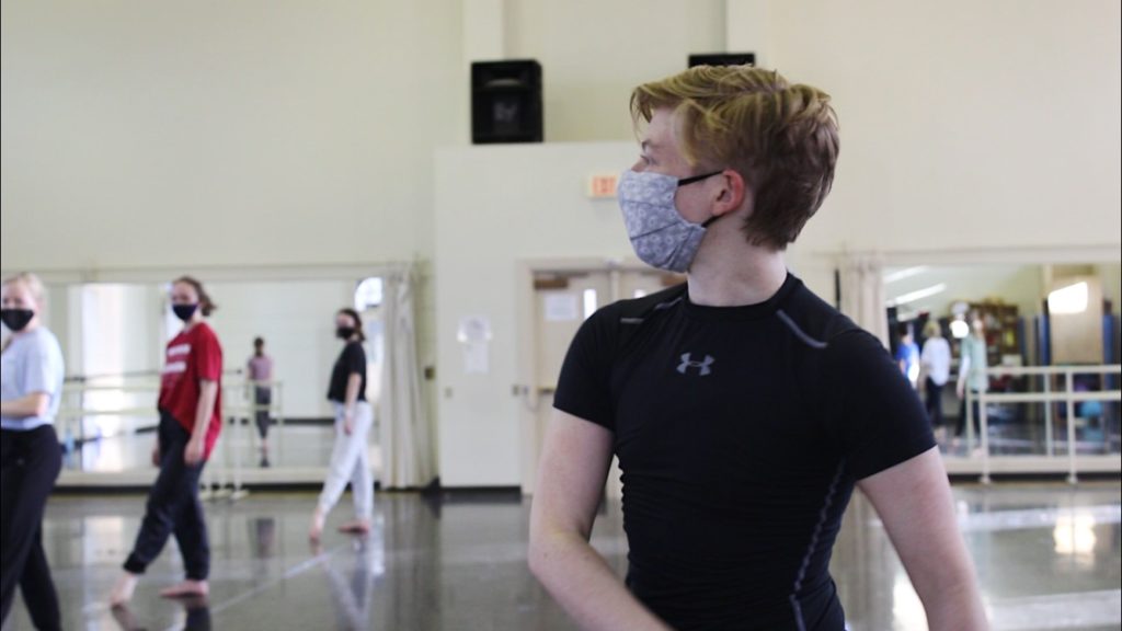 A dance teacher wearing a mask teaches in a classroom of dance students.