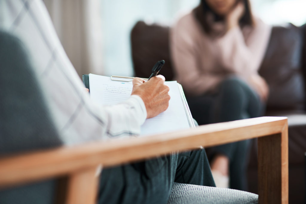 A therapist sitting in a chair writes in their notepad while listening to a patient during a therapy session.