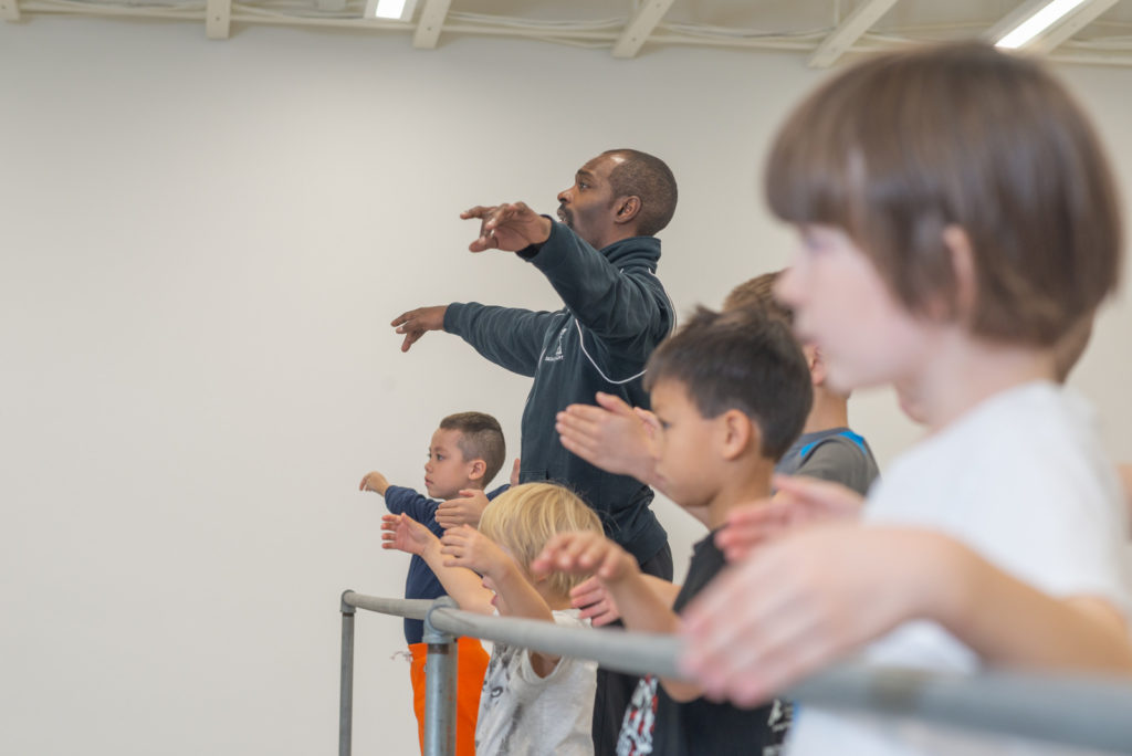 Jacob Ashley Brooks EL is shown in profile as he demonstrates port de bras, his arms in allongé second. Young male ballet students stand at barre imitating him.