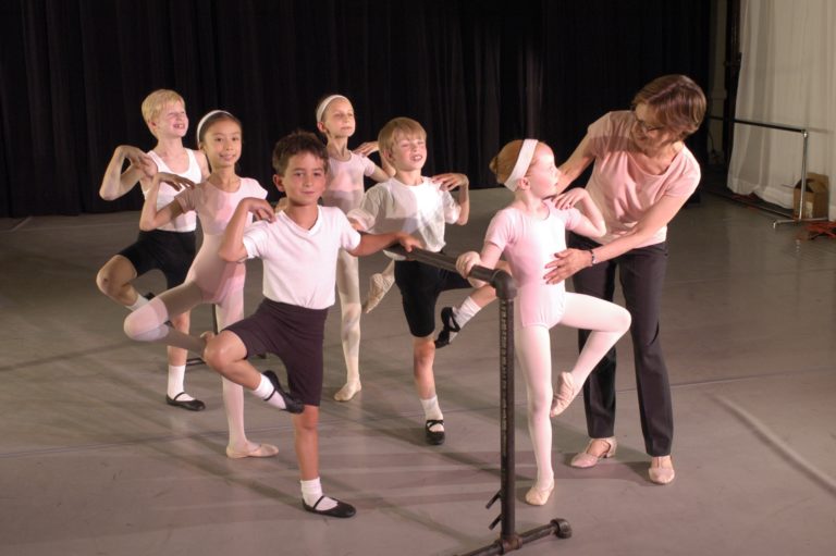 Image of young ballet students at the barre in a ballet class with one student being supported by educator Diana Byer