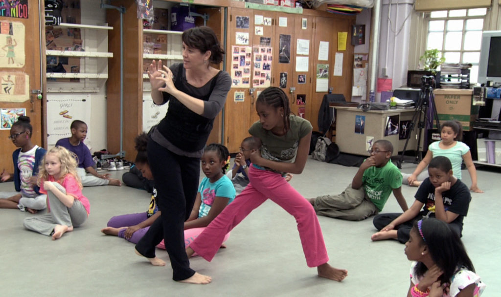 A woman dance teacher teaches a classroom of young dance students.