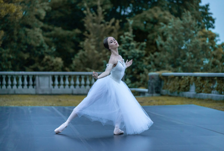 A ballerina in a long, white tutu dances on a stage outdoors.