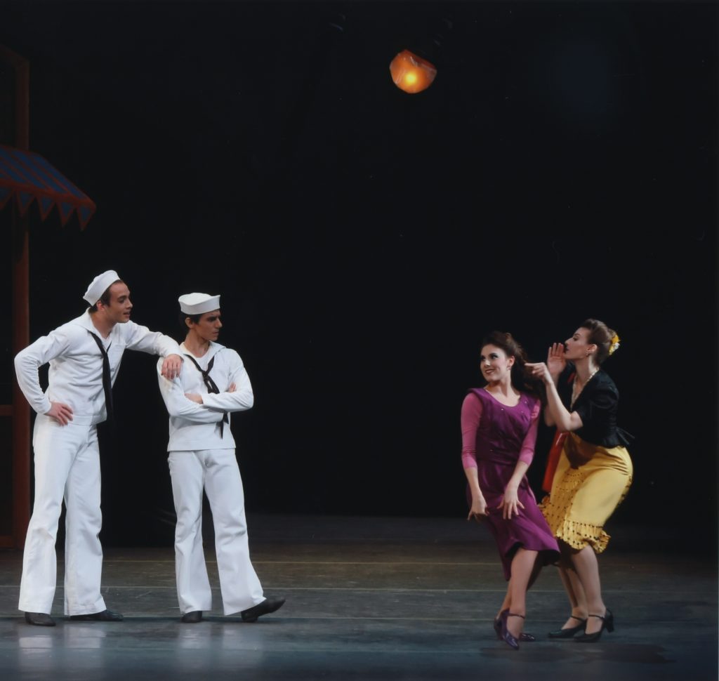 Four dancers, two men in sailor costumes and two women in colorful dressed, perform on stage.