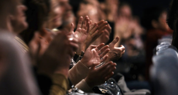 View down a row of people sitting in an audience of people clapping their hands in appreciation of a performance