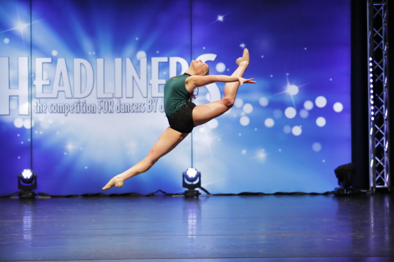 A young dancer photographed mid-leap with her head and torso arched back toward her left leg is back attitude.