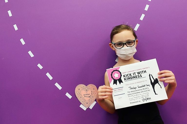 A dance student wearing a mask poses holding a certificate in front of a purple wall with a white-outline heart on it.