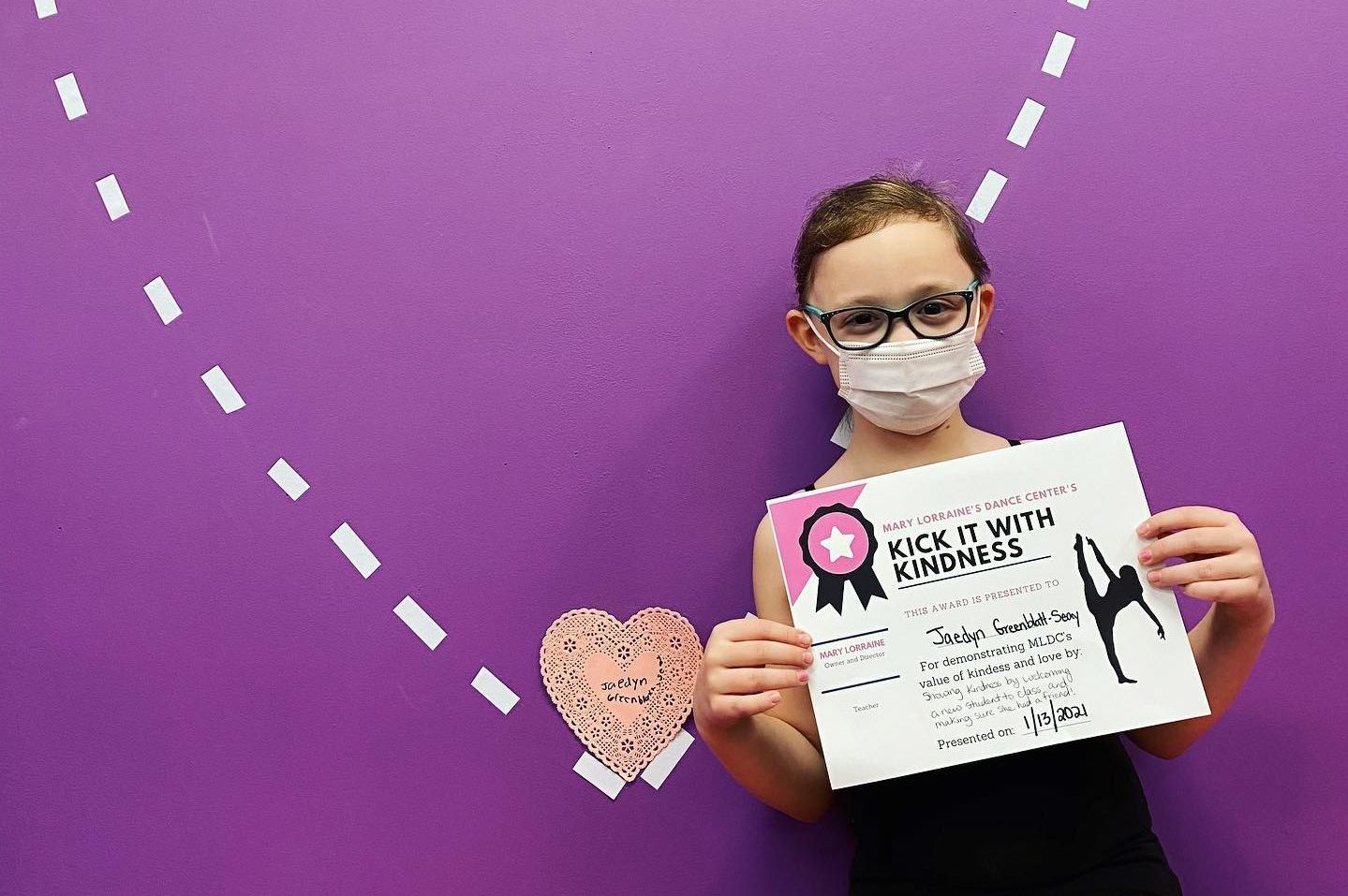 A dance student wearing a mask poses holding a certificate in front of a purple wall with a white-outline heart on it.
