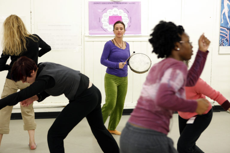 A dance teacher holding a drum teaches a dance class full of moving dancers.