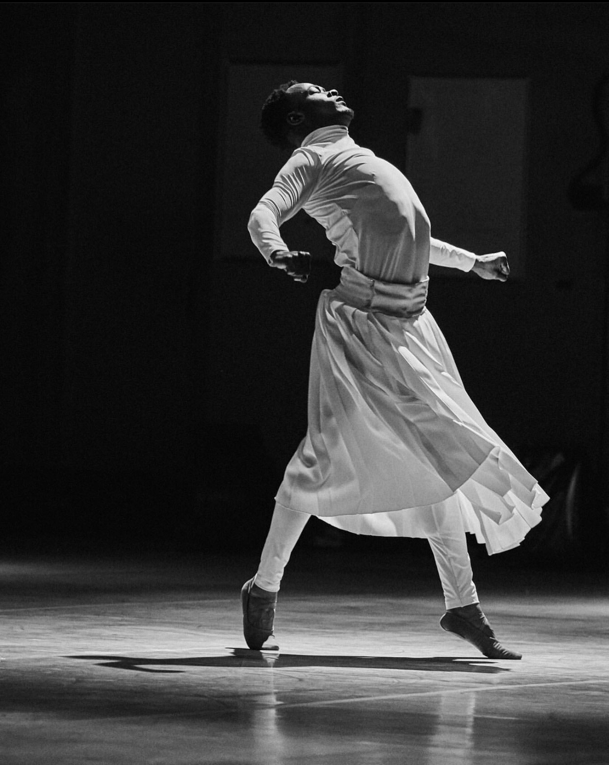 A black and white image of a male dancer in a white top and long skirt performing on stage.