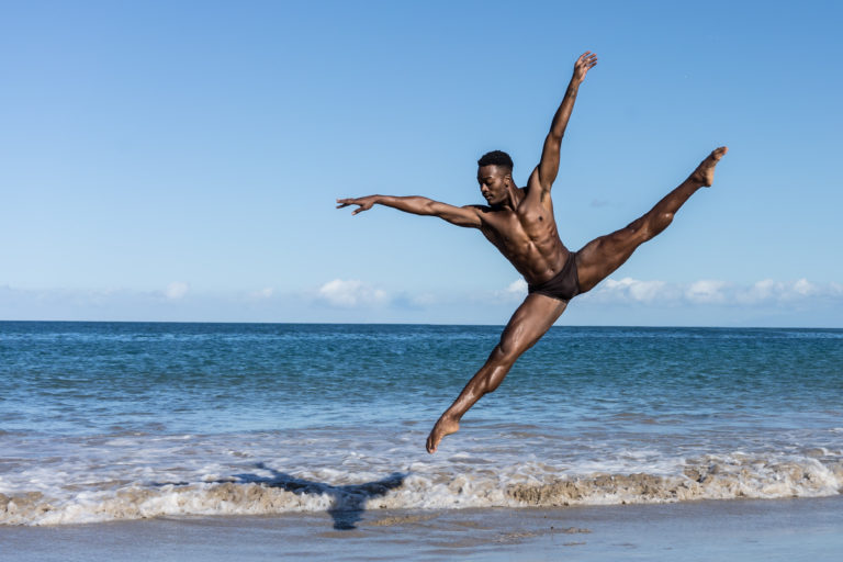 A male dancer does a tilt jump on the beach in front of the ocean.