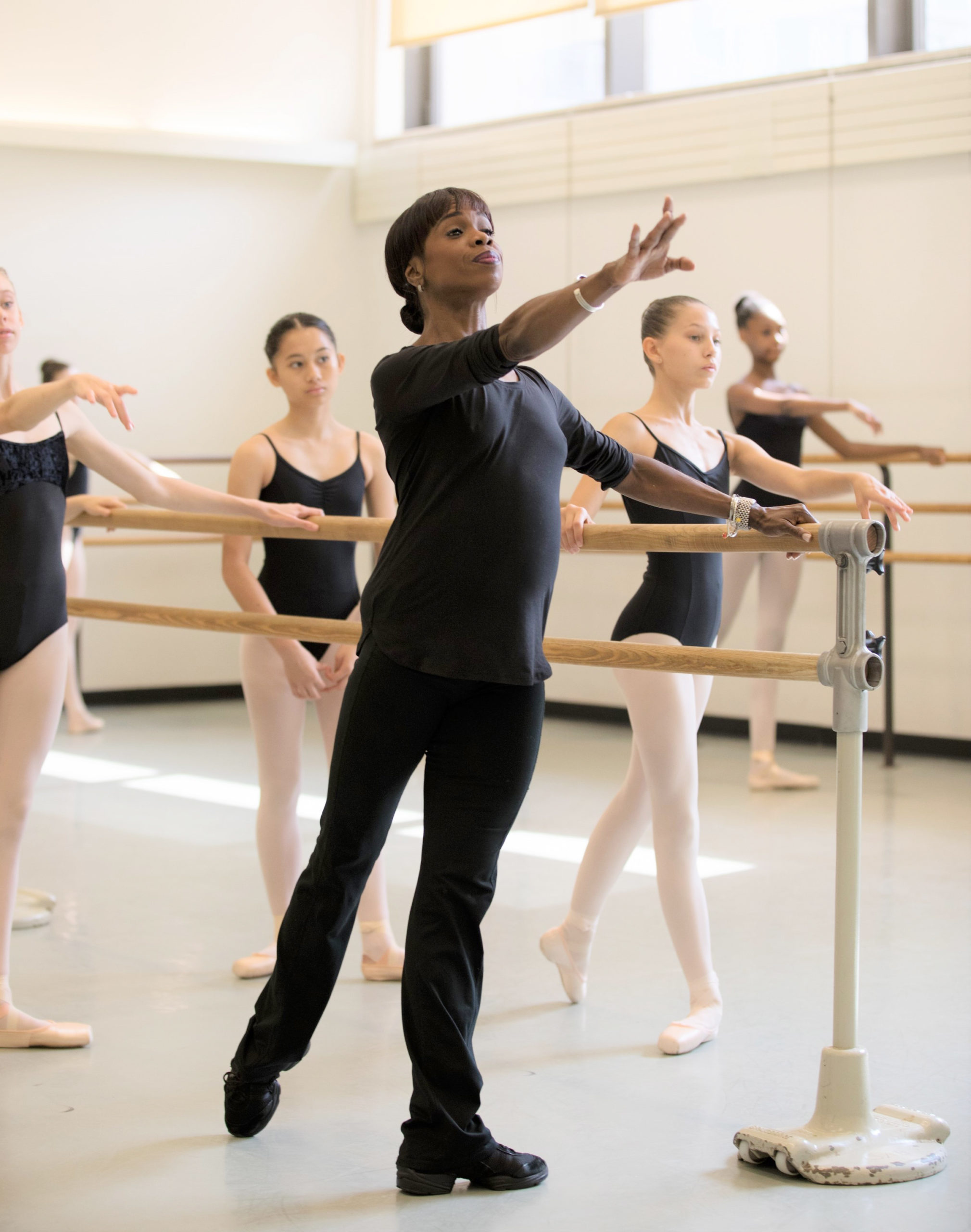 Andrea Long, a tall Black woman wearing black sweats, dance sneakers, and shirt, stands at barre, demonstrating a tendu back, working arm extended forward in arabesque. Students in pink tights and black leotards watch and follow along.