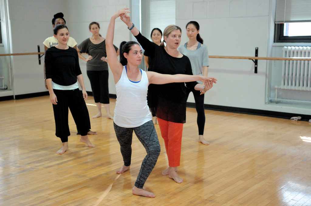 Deborah Damast, wearing a long-sleeved black top and orange pants, instructs a graduate dance student as five other students watch in the background.