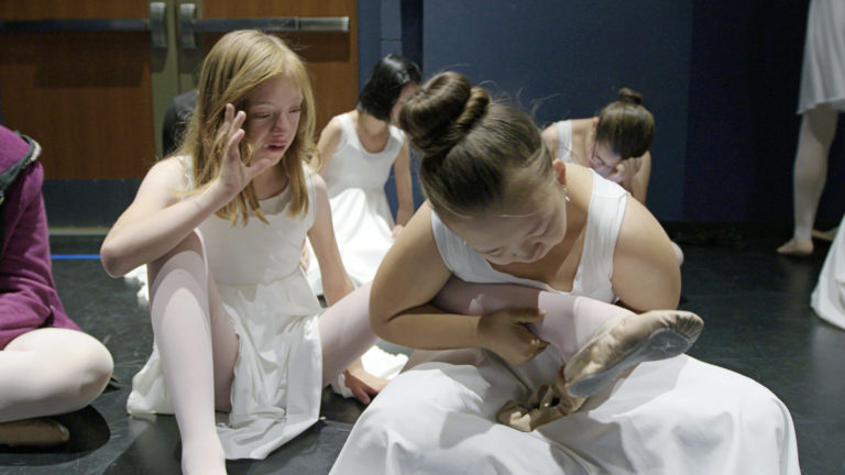 Young student helping her peer with putting on ballet shoes