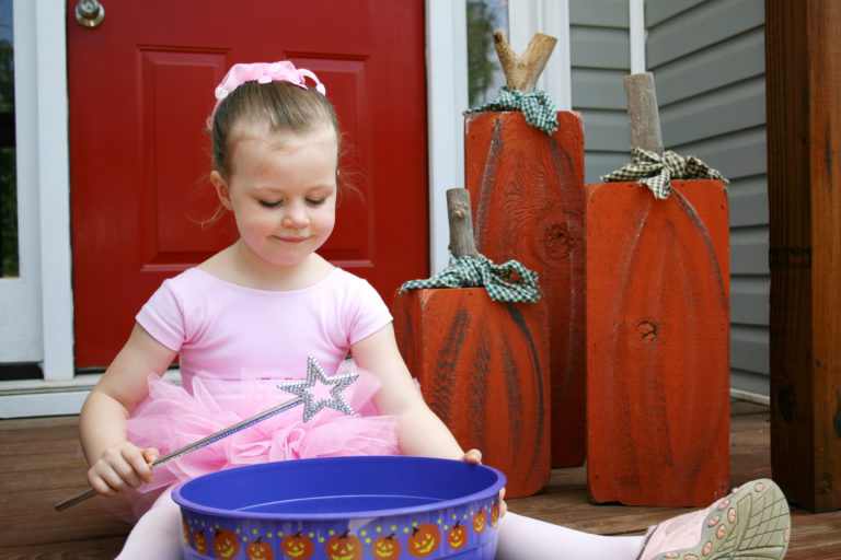 Young ballerina seated holding a wand, bucket with halloween pumpkins, and decorated pumpkins behind her