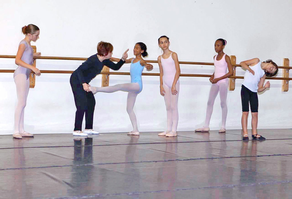 A ballet teacher in black clothing corrects a young female ballet student at the barre, holding the student's leg devant and pointing her finger as she gives the correction. The young student wears a light blue leotard, pink tights and ballet shoes. Three other young students watch in light pink leotards, tights and shoes.