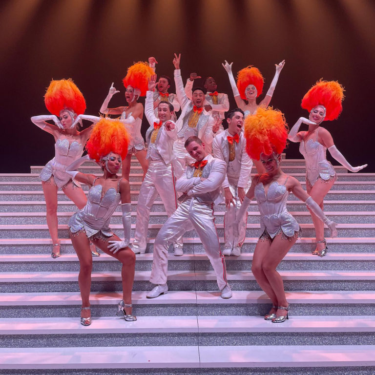 Group of dancers with white costumes and orange/red feather wigs