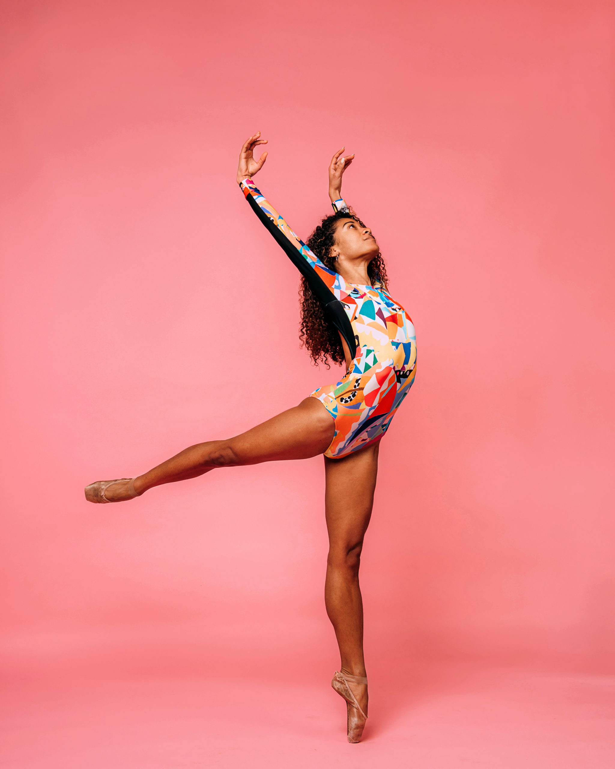 Princess Reid poses in a low arabesque, arms lifted above her head and head turned upward confidently, in a multicolored long-sleeve leotard. Her long brown hair is down, and she dances on pointe in front of a rosy pink backdrop.