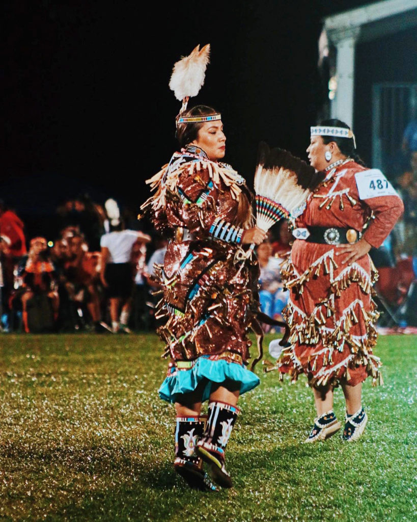 Kendorina Redhouse performs a Native American dance in traditional clothing, her long brown dress adorned with brightly colored wooden beads and embroidery. She wears a feather headdress and moccasins and dances with a fan made of feathers. Onlookers watch as she dances in the grass during the evening. Another dancer in the competition dances behind her, to the right.