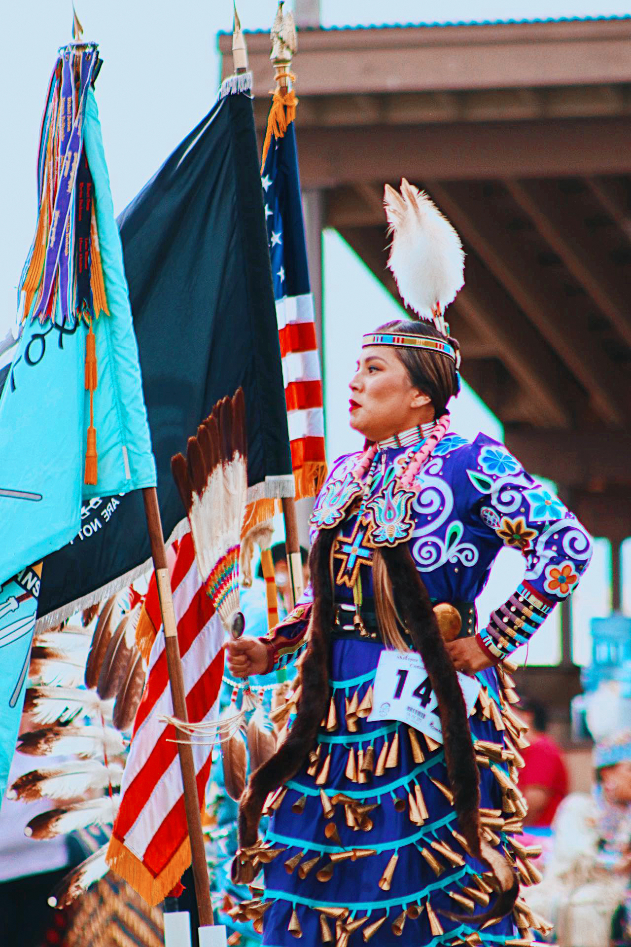 Kendorina Redhouse stands next to a Native American flag and the US flag in traditional clothing, her long purple dress adorned with brightly colored wooden beads and embroidery. She wears a feather headdress and moccasins and holds with a fan made of feathers.