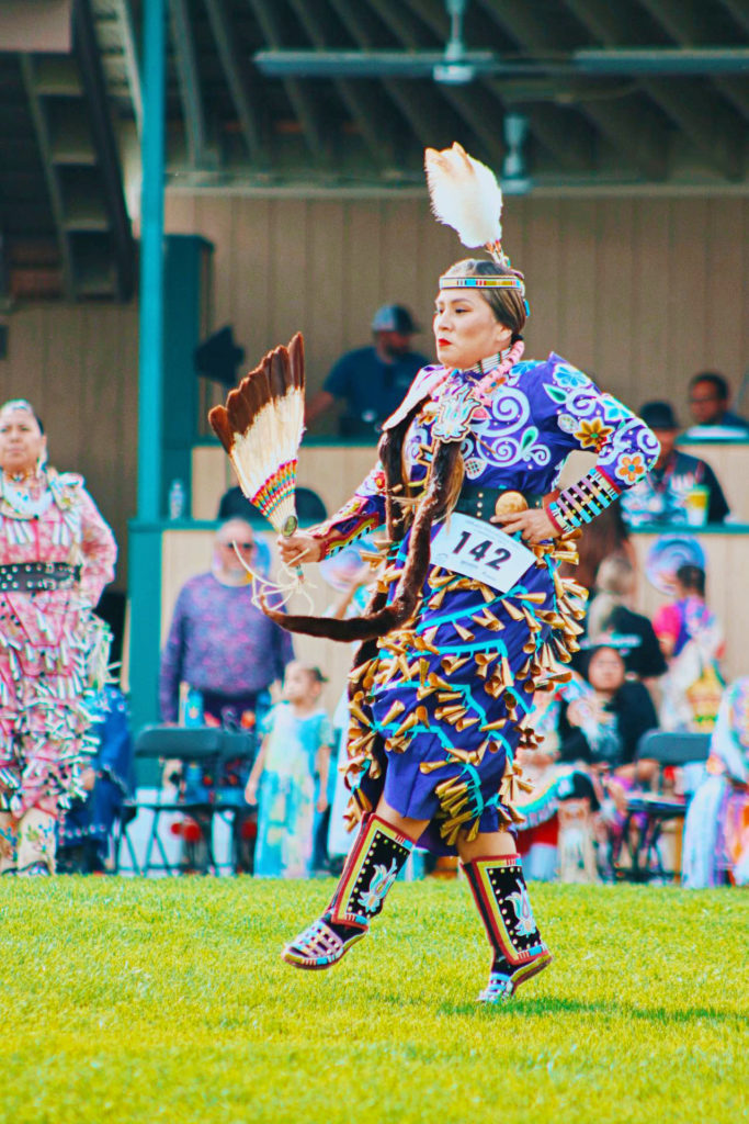 Kendorina Redhouse performs a Native American dance in traditional clothing, her long purple dress adorned with brightly colored wooden beads and embroidery. She wears a feather headdress and moccasins and dances with a fan made of feathers. Onlookers watch as she dances in the grass on a sunny day.