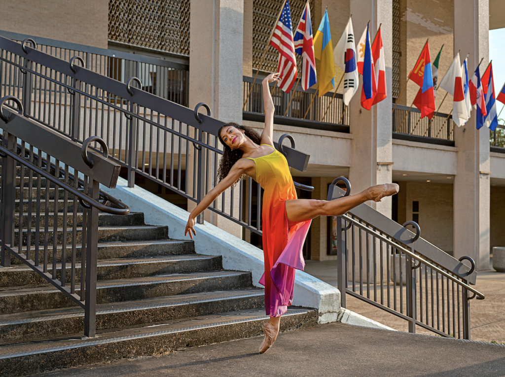 Princess Reid poses in a bright yellow, red and purple dance dress. She is on pointe, extending her right leg devant and does a cambre back with arms extended in allonge. She smiles at the camera. She dances outside of an embassy building by a staircase, with flags in the background.