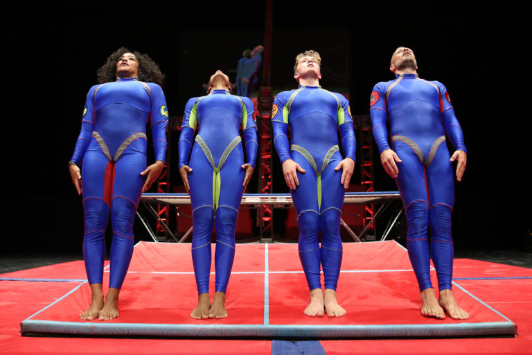 Image of 4 dancers wearing blue jumpsuits learning backwards into a fall with cushioning underneath them.