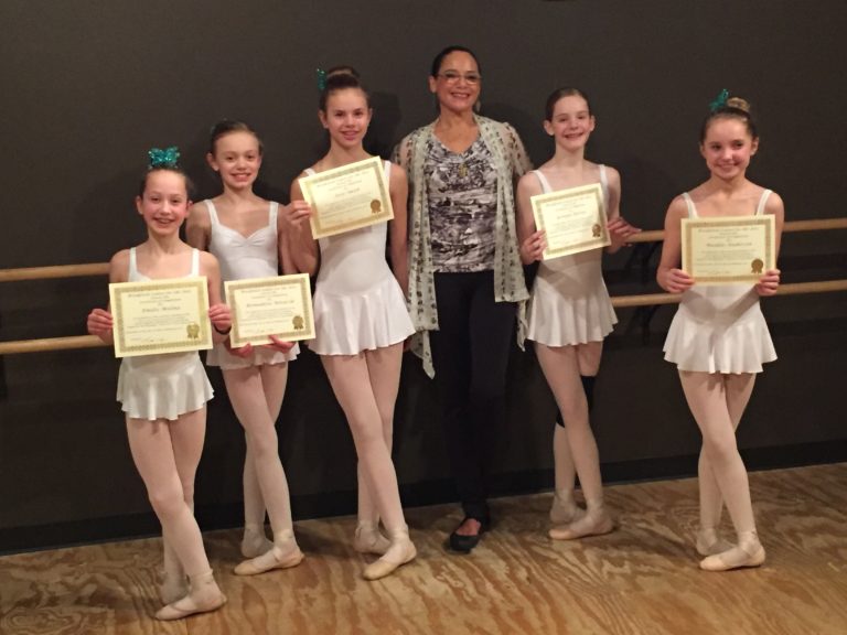 Dance teacher posing with her ballet students who are all holding up certificates.