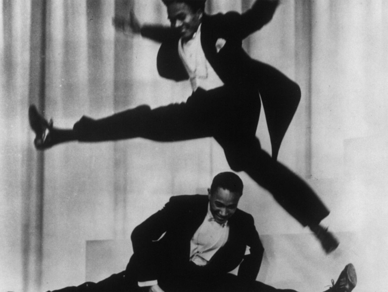 A black and white photograph. Harold Nicholas jumps over his brother, Fayard. They wear tuxedos and tap shoes and perform in front of a stage curtain.