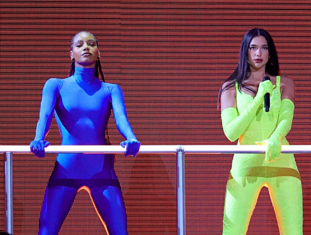 Demi Rox (left) wears a bright blue longsleeve spandex suit and poses confidently onstage, her hands gripping a metal bar in front of her. Dua Lipa (right) wears a highlighter yellow spandex suit and sings into a microphone.