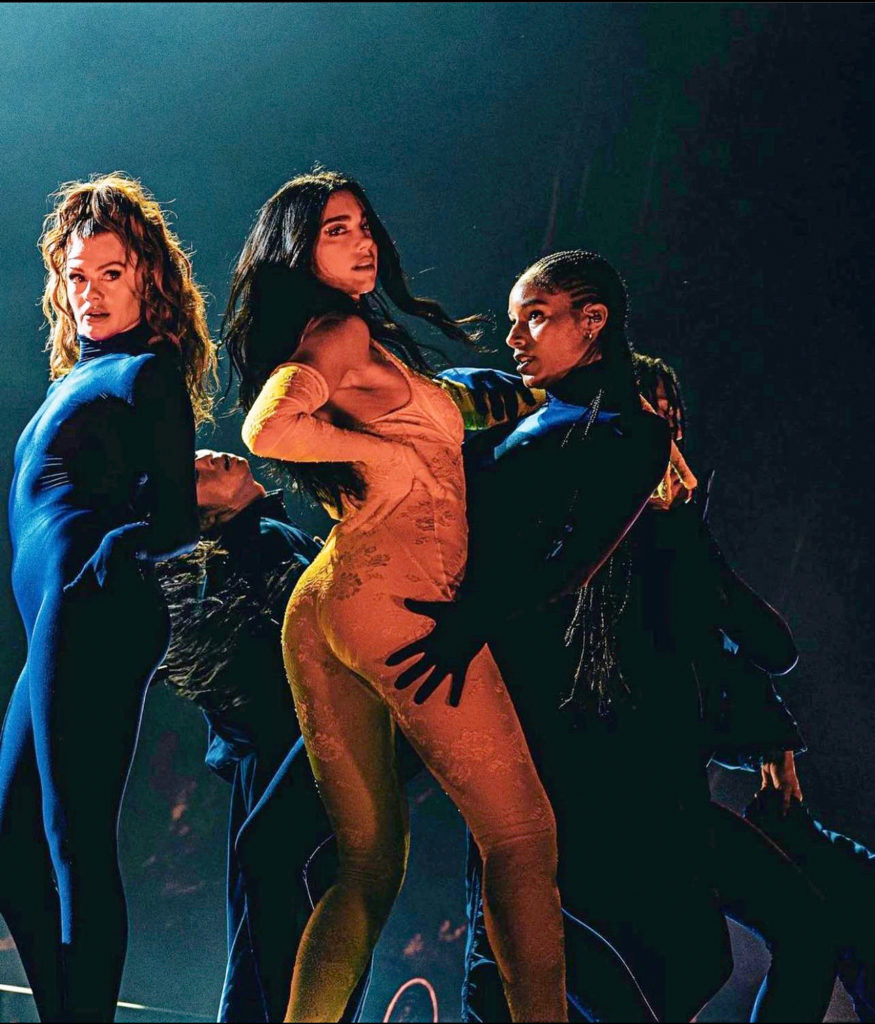 Dua Lipa wears a tan textured longsleeve spandex suit. Backup dancers beside and behind her wear longsleeved black spandex suits and pose around her. They all have their long hair down.