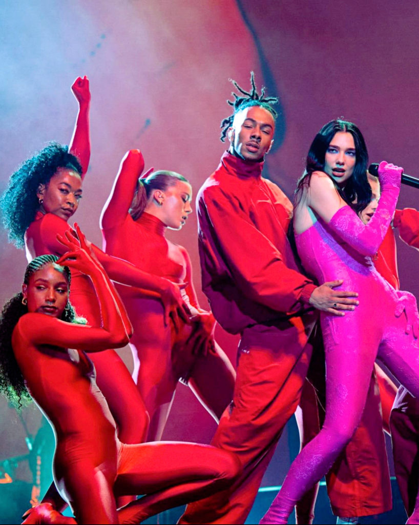 3 female and 1 male backup dancer wear long-sleeved bright red spandex suits with their hair down onstage as they perform with Dua Lipa, who wears a hot pink spandex suit.