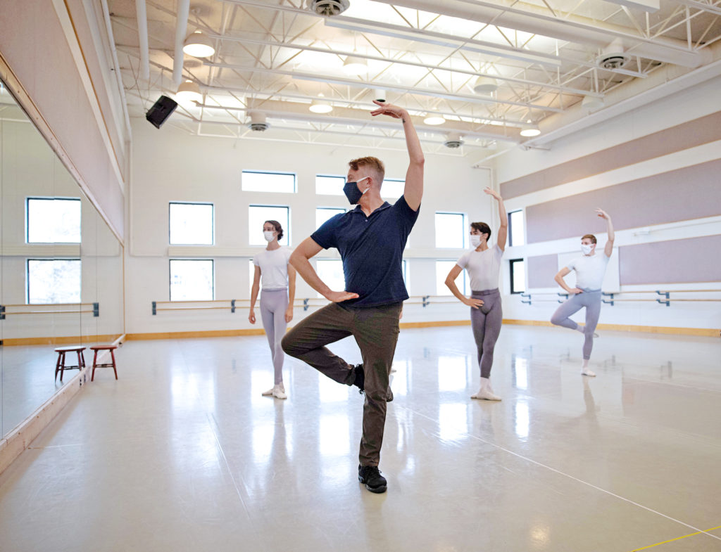 Chris Alloways-Ramsey teaches ballet class in a brightly lit studio. He wears all black and demonstrates retiré as young male ballet students watch.