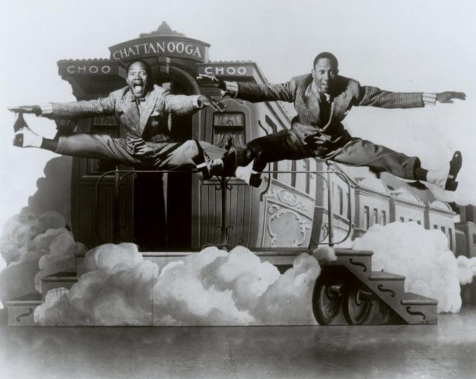 A black and white photograph. The Nicolas Brothers split jump together in front of a steaming train.